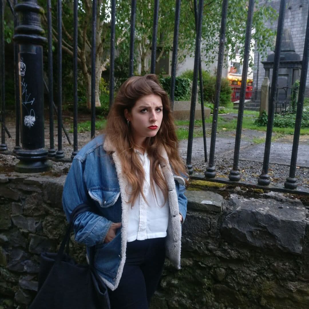 A woman with long brown hair poses in front of a graveyard.